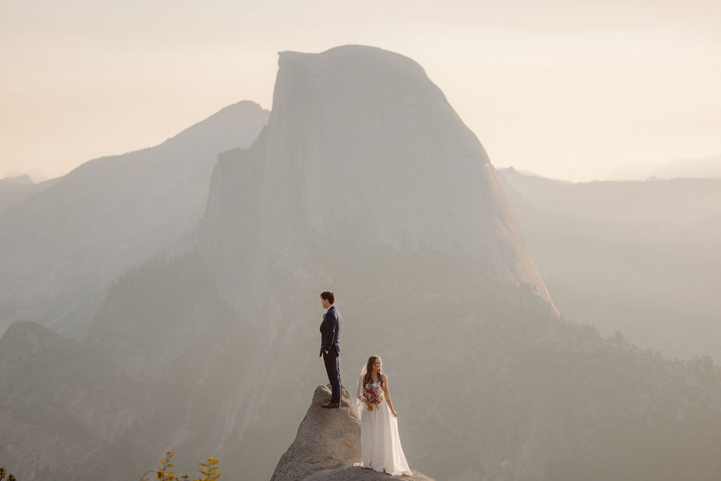 A bride in a white dress stands beside a groom in a suit on a rocky ledge, with mountains and trees in the background for a wedding at glacier point