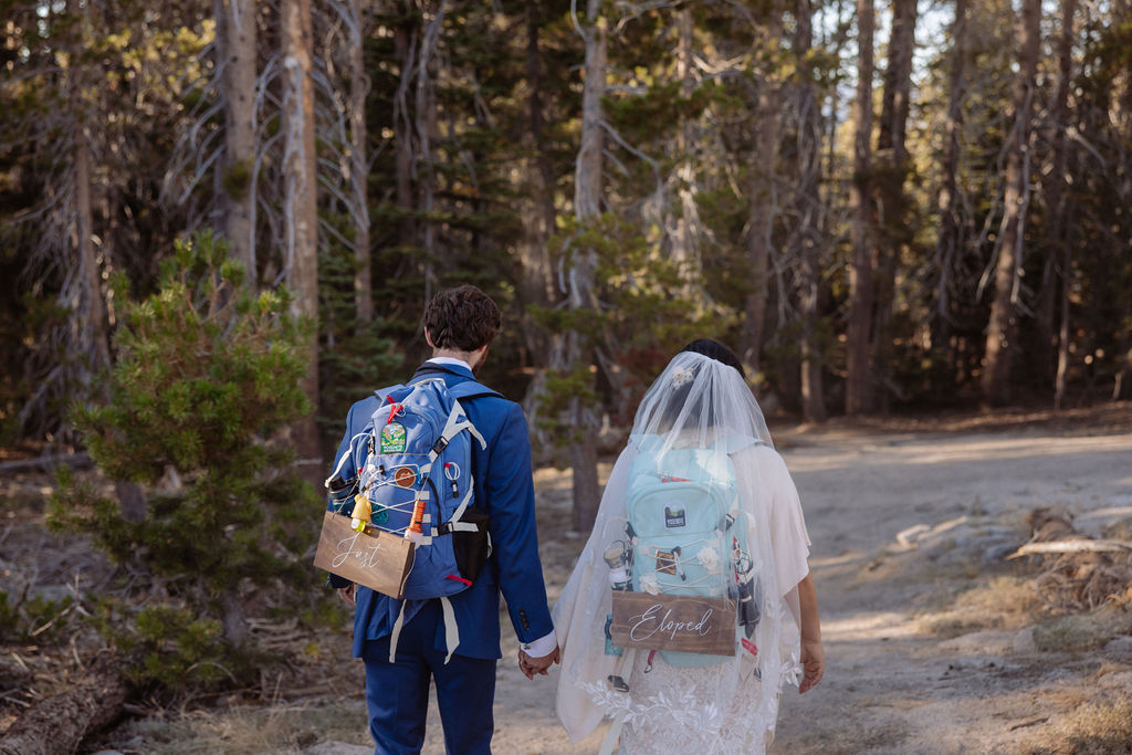 A bride and groom wearing backpacks and holding hands walk on a forest trail, with a "Just Married" sign on the bride's backpack