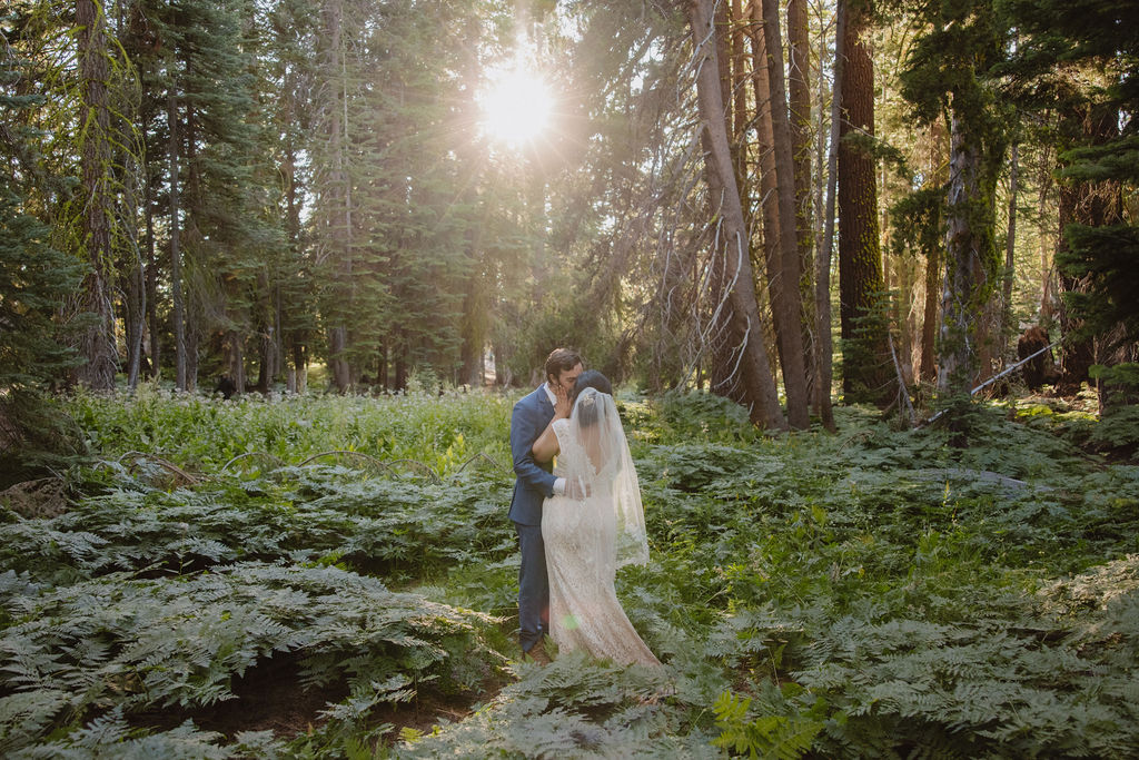 A bride and groom walk hand in hand through a forest, surrounded by tall trees and green vegetation.