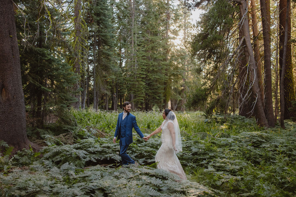 A bride and groom walk hand in hand through a forest, surrounded by tall trees and green vegetation.