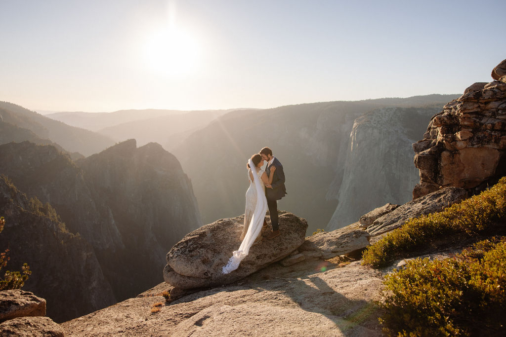 A bride and groom stand on a large rock at the edge of a cliff, facing each other and holding papers, with mountains in the background.
