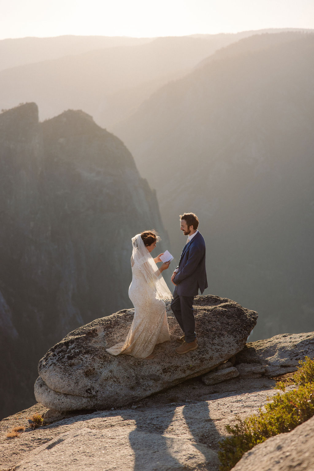 A bride and groom stand together on a rocky cliff at sunset with mountains in the background for a wedding at Yosemite