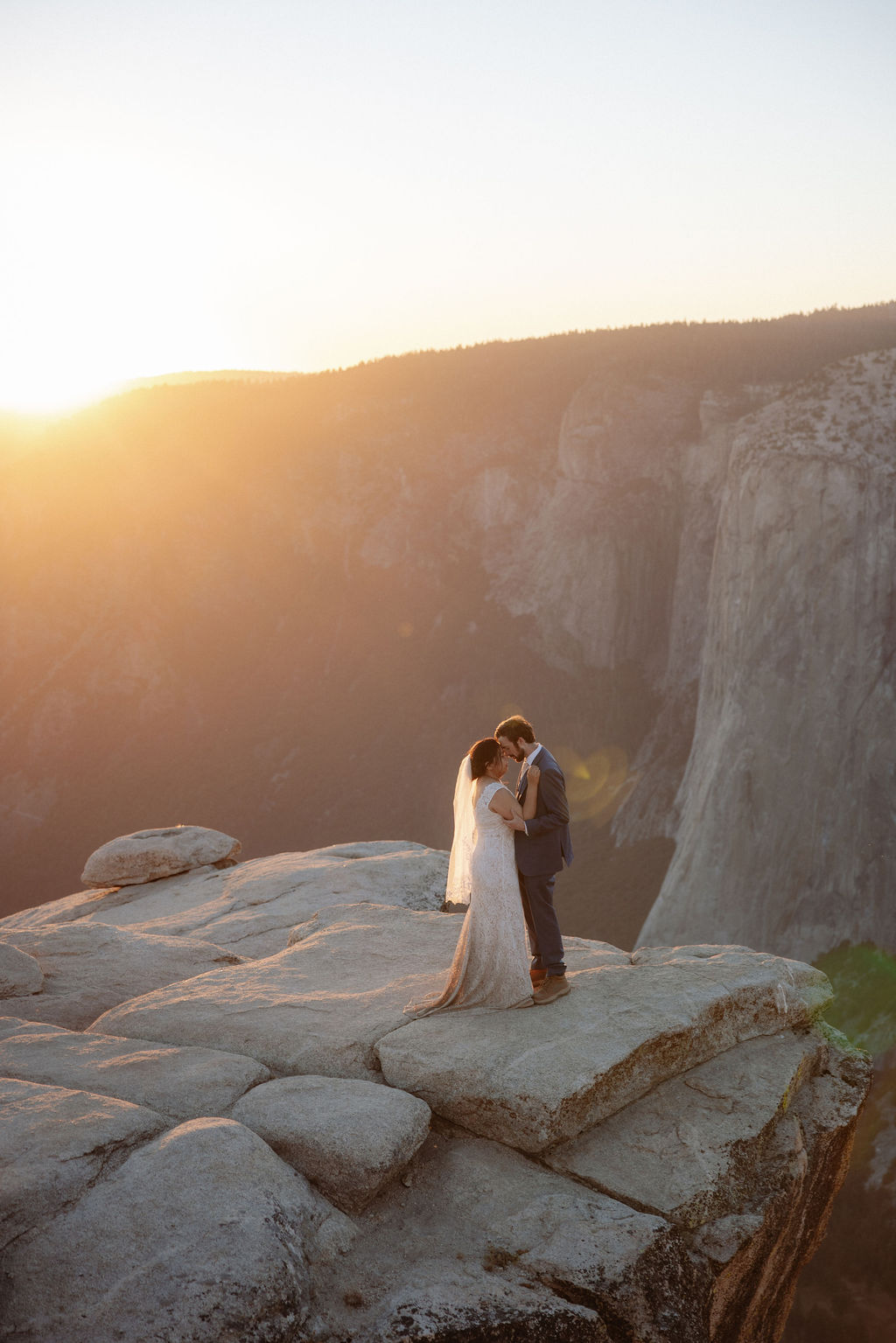 A bride and groom stand together on a rocky cliff at sunset with mountains in the background for a wedding at Yosemite