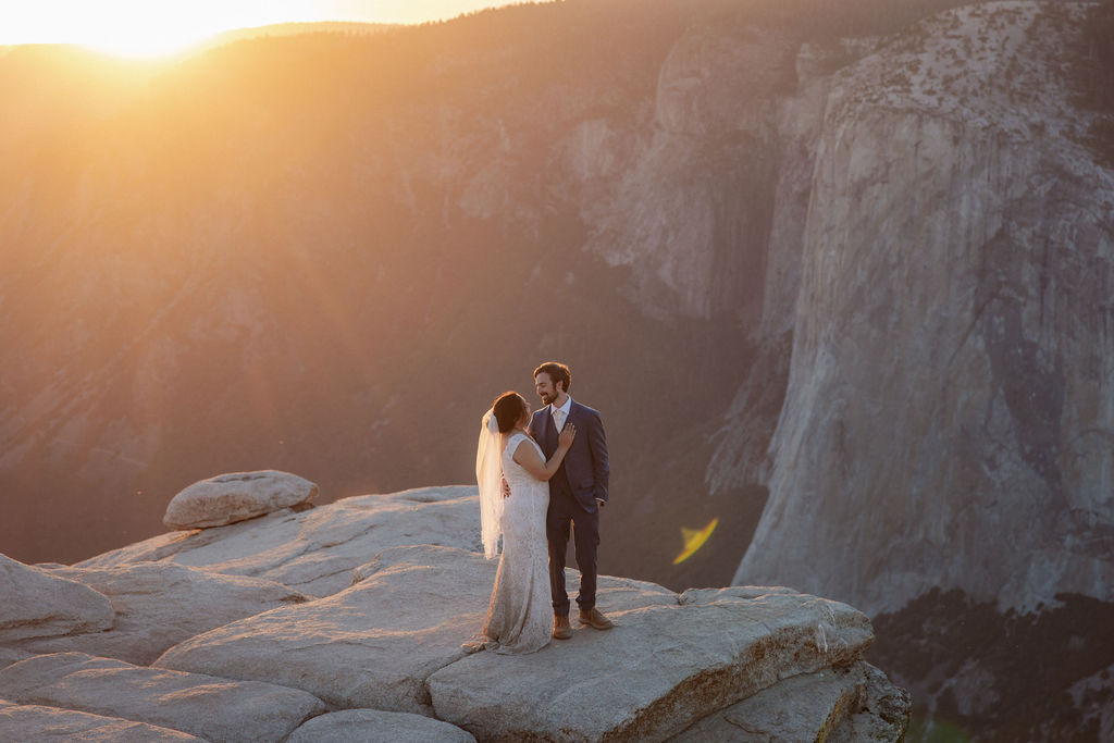 A bride and groom stand together on a rocky cliff at sunset with mountains in the background for a wedding at Yosemite