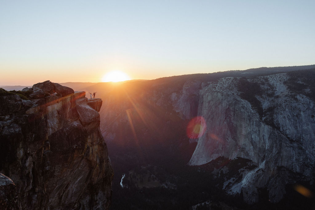 A person stands on the edge of a rocky cliff at sunset, overlooking a valley with distant mountains and a bright sun on the horizon.