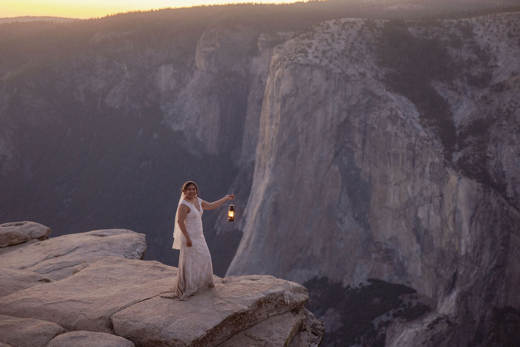 A bride and groom stand on a rocky cliff edge overlooking a deep canyon at sunset, with distant mountains in the background.