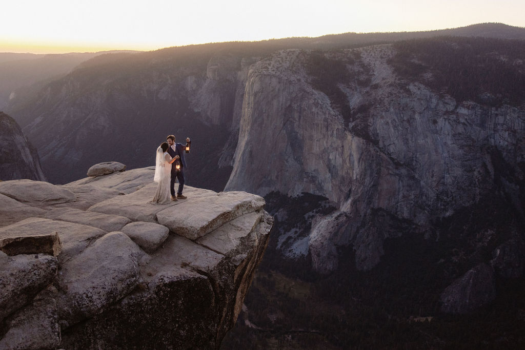 A bride and groom stand on a rocky cliff edge overlooking a deep canyon at sunset, with distant mountains in the background.