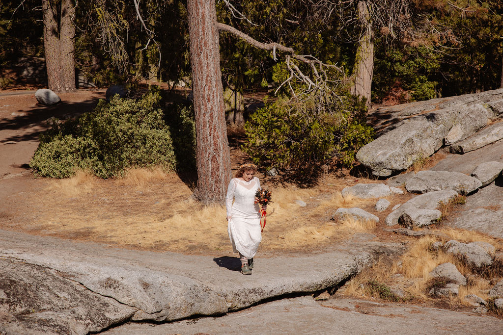 A person in a white dress holding a bouquet walks on a rocky path in a forested area with dry grass and tall trees.