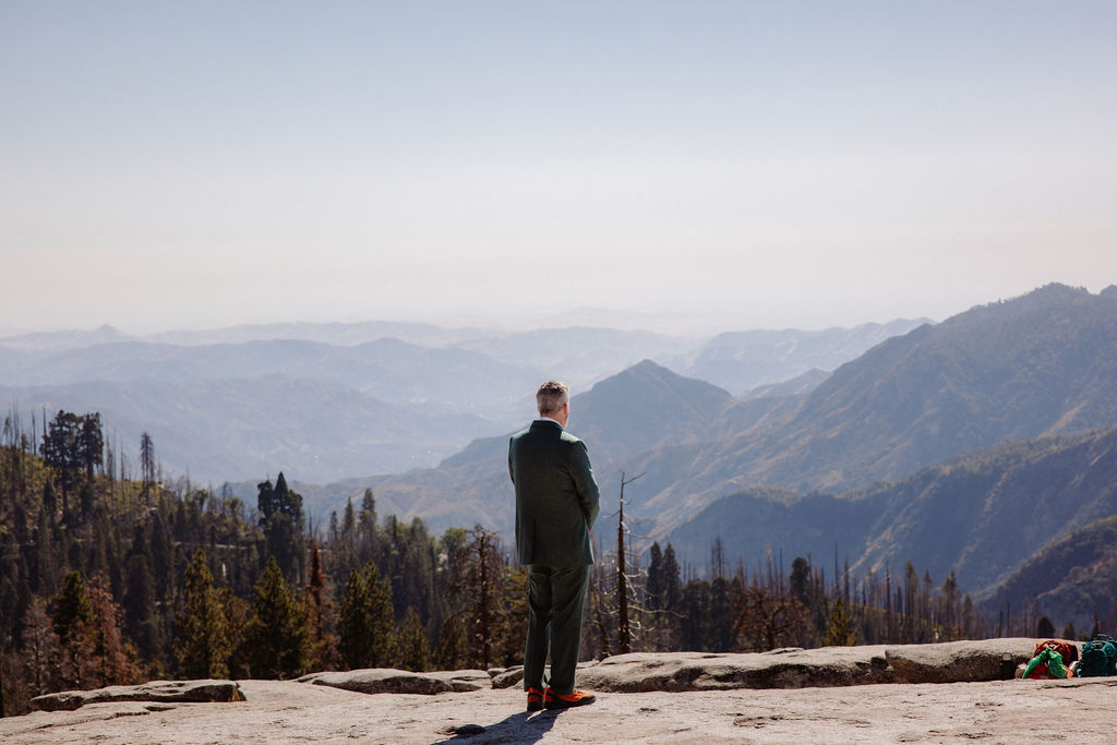 A person in a suit stands on a rocky ledge overlooking distant forested mountains under a clear sky.