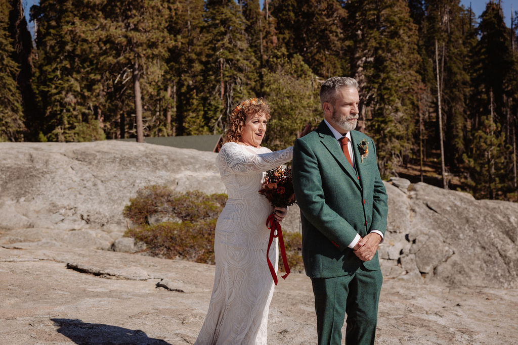 A bride in a white dress stands behind a groom in a green suit during an outdoor first look moment in a rocky, forested area.
