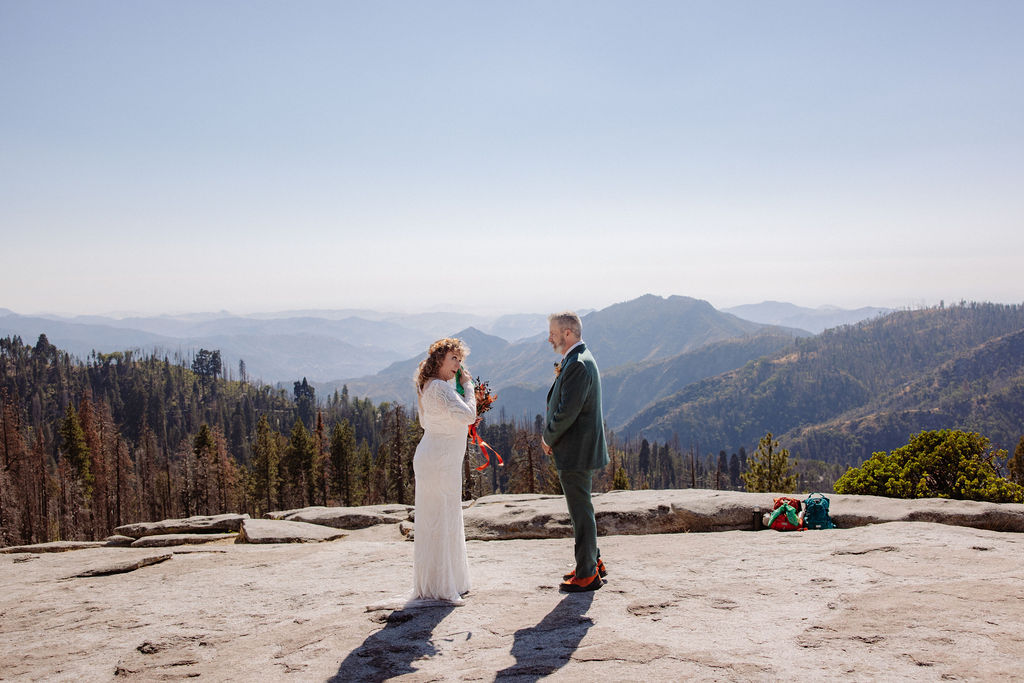 A bride and groom stand facing each other on a rocky overlook with mountains and trees in the background on a clear day.
