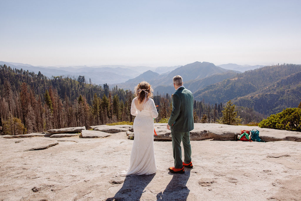A bride and groom stand facing each other on a rocky overlook with mountains and trees in the background on a clear day.
