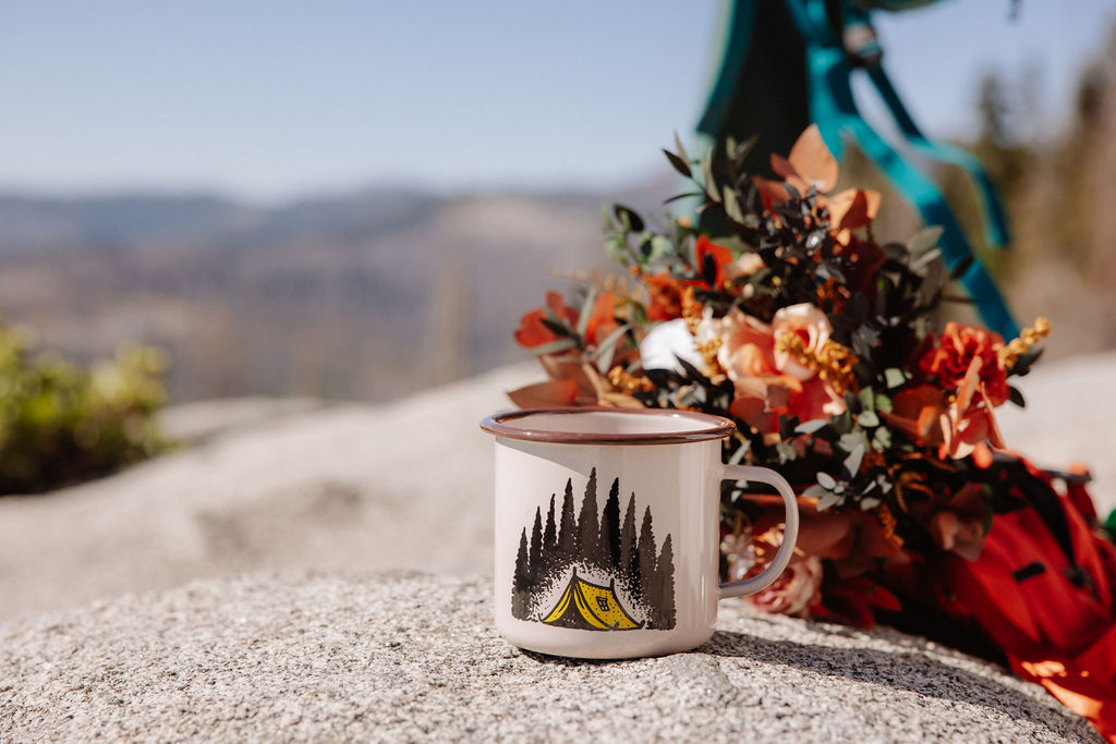 A white enamel mug with a tent and trees design sits on a rock beside a bouquet of autumn-colored flowers, with blurred mountains in the background.