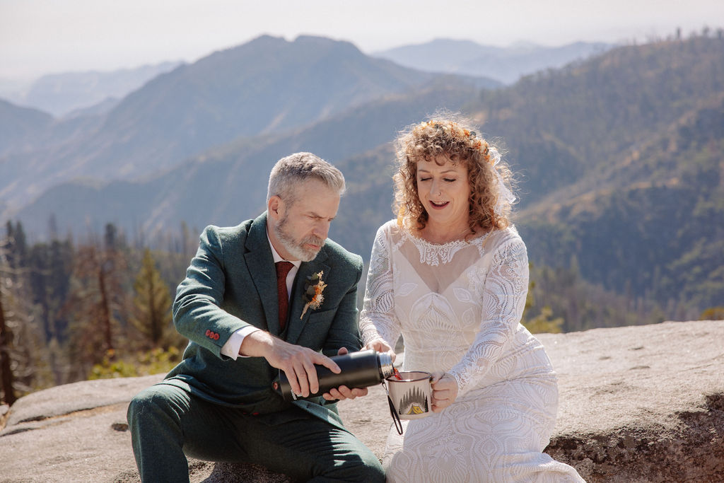 A bride and groom in wedding attire sit outdoors on a stone ledge, holding metal mugs and smiling at each other with mountains in the background taken by a sequoia elopement photographer
