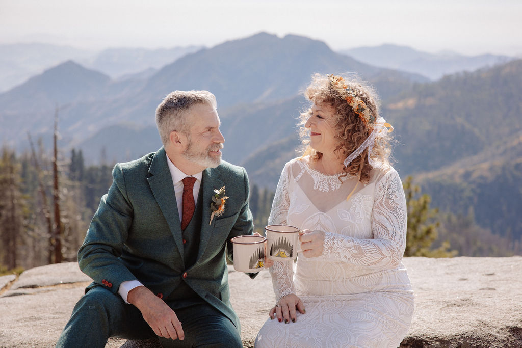 A bride and groom in wedding attire sit outdoors on a stone ledge, holding metal mugs and smiling at each other with mountains in the background taken by a sequoia elopement photographer