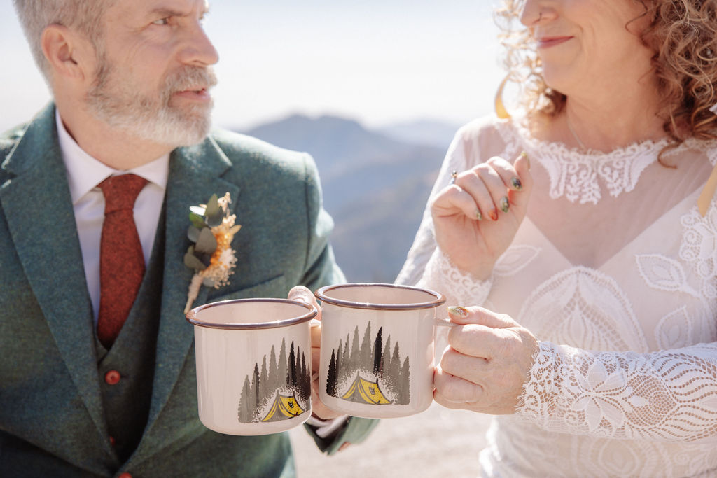 A bride and groom in wedding attire sit outdoors on a stone ledge, holding metal mugs and smiling at each other with mountains in the background taken by a sequoia elopement photographer