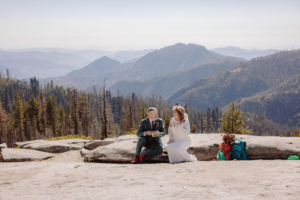 A bride and groom in wedding attire sit outdoors on a stone ledge, holding metal mugs and smiling at each other with mountains in the background taken by a sequoia elopement photographer