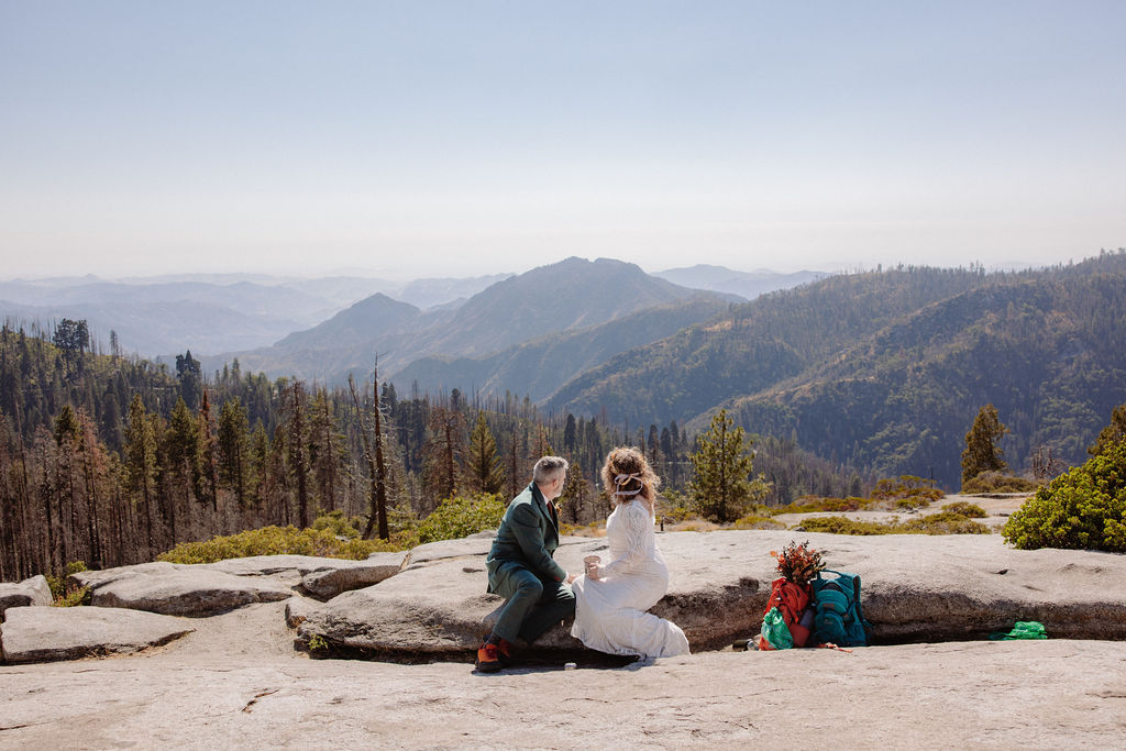 A bride and groom in wedding attire sit outdoors on a stone ledge, holding metal mugs and smiling at each other with mountains in the background taken by a sequoia elopement photographer