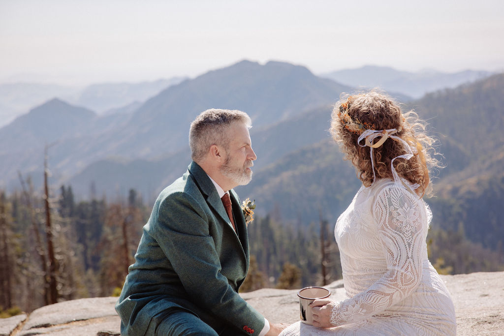 A bride and groom in wedding attire sit outdoors on a stone ledge, holding metal mugs and smiling at each other with mountains in the background taken by a sequoia elopement photographer