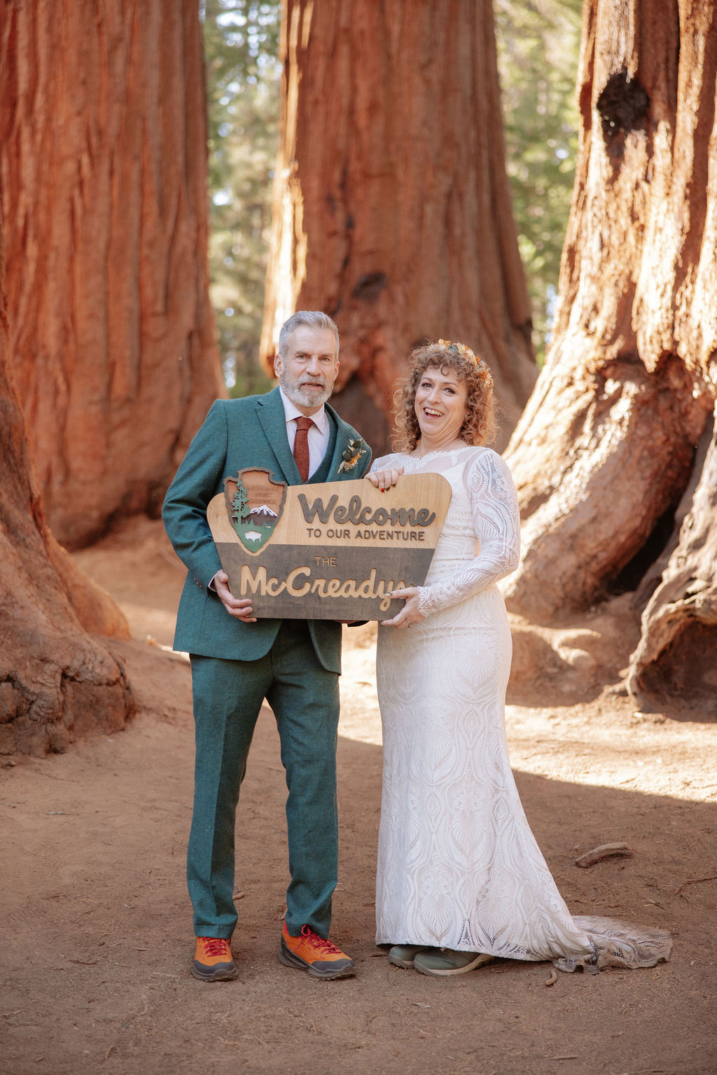 A man in a green suit and a woman in a white dress stand outdoors among giant trees, holding a wooden "Welcome McCreadys" sign.