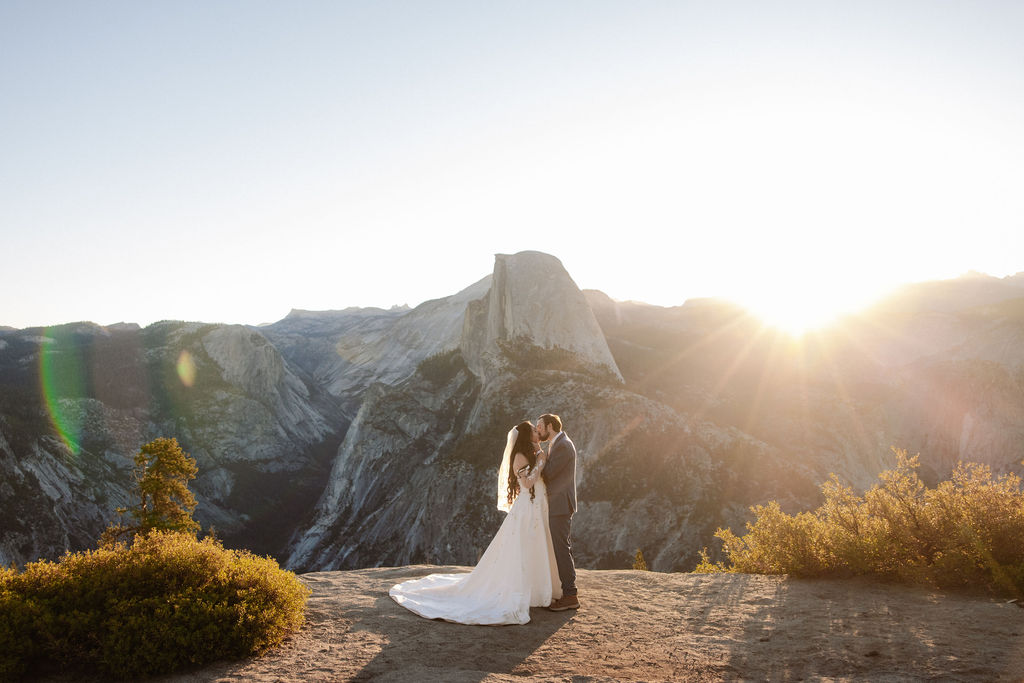 A couple in wedding attire stands on a rocky ledge overlooking a mountain landscape with Half Dome in the background at sunrise for a wedding at Yosemite
