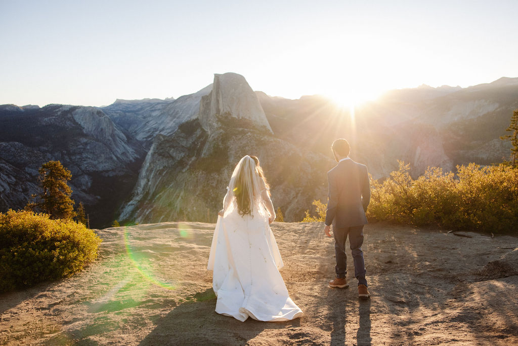 A couple in wedding attire stands on a rocky ledge overlooking a mountain landscape with Half Dome in the background at sunrise for a wedding at Yosemite