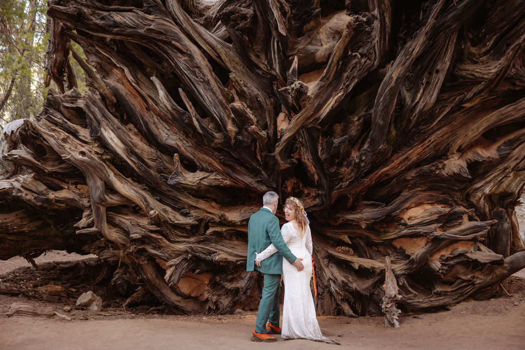 A couple dressed in formal wedding attire stands together at the base of large redwood trees in a forest taken by sequoia elopement photographer