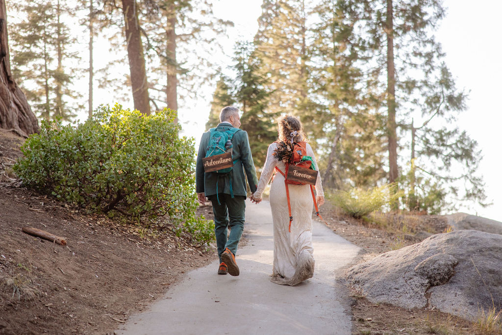 A couple dressed in wedding attire, holding hands and walking on a forest path with backpacks that say "Just Married