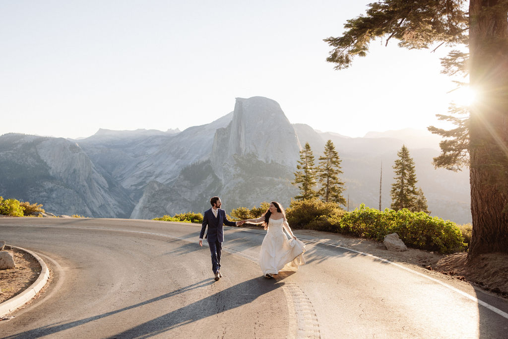 A couple in wedding attire walks hand in hand on a curved road with mountains and trees in the background at sunset.
