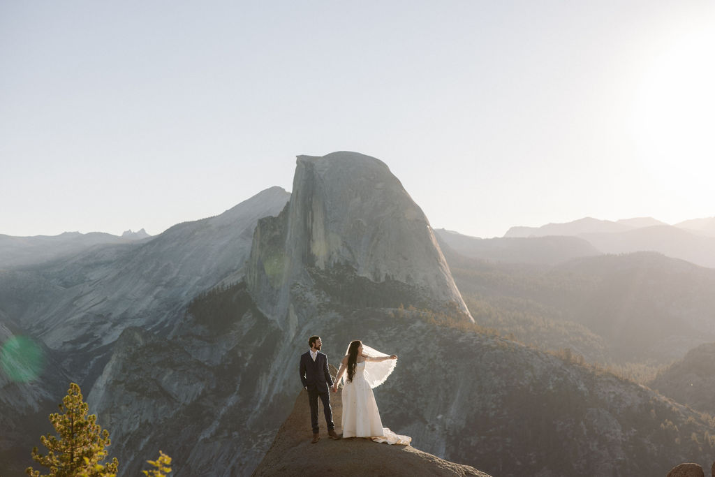 A couple in wedding attire stands on a rocky ledge overlooking a mountain landscape with Half Dome in the background at sunrise for a wedding at Yosemite