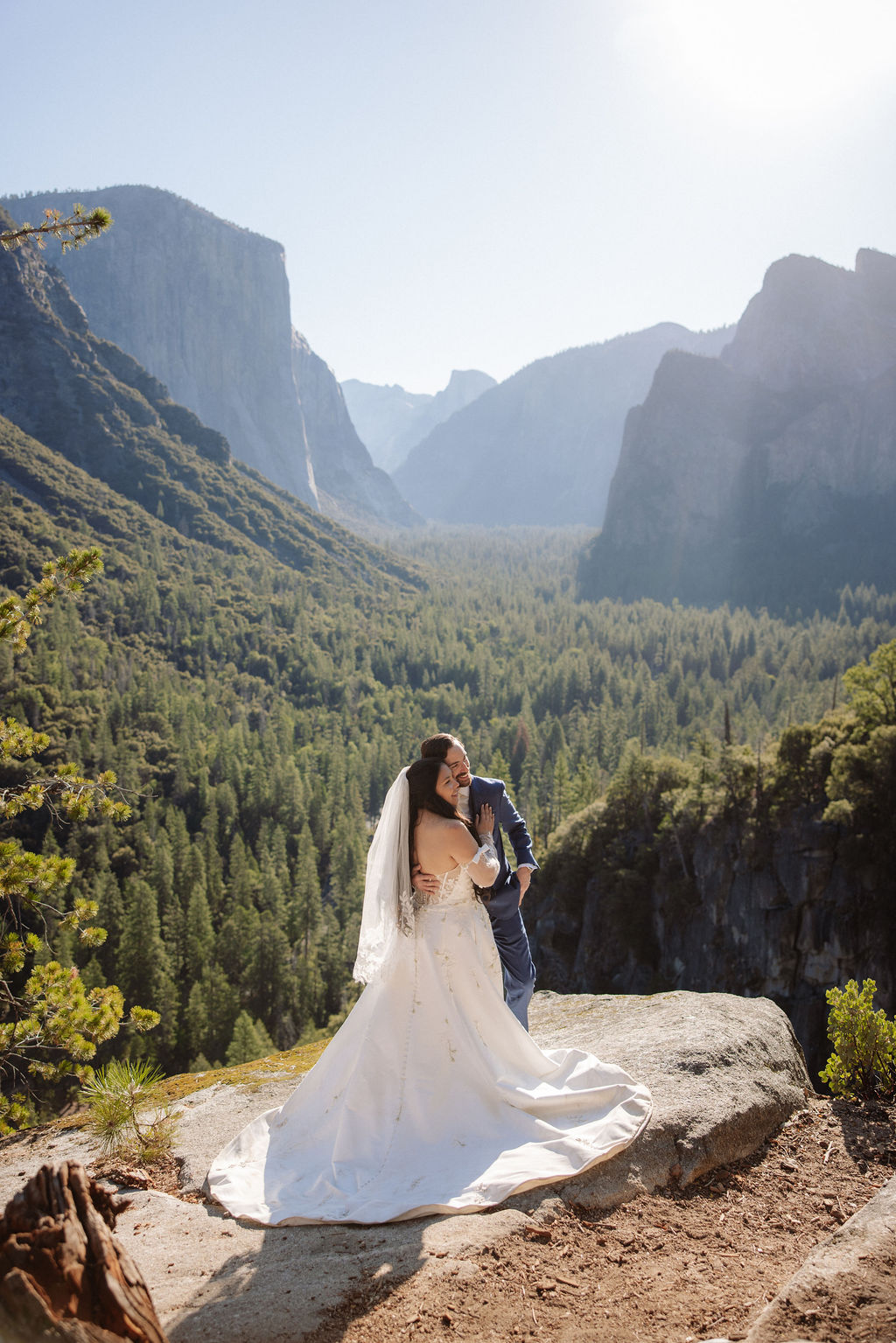 A bride and groom stand together on a rocky ledge overlooking a forested valley and mountains under a clear sky.