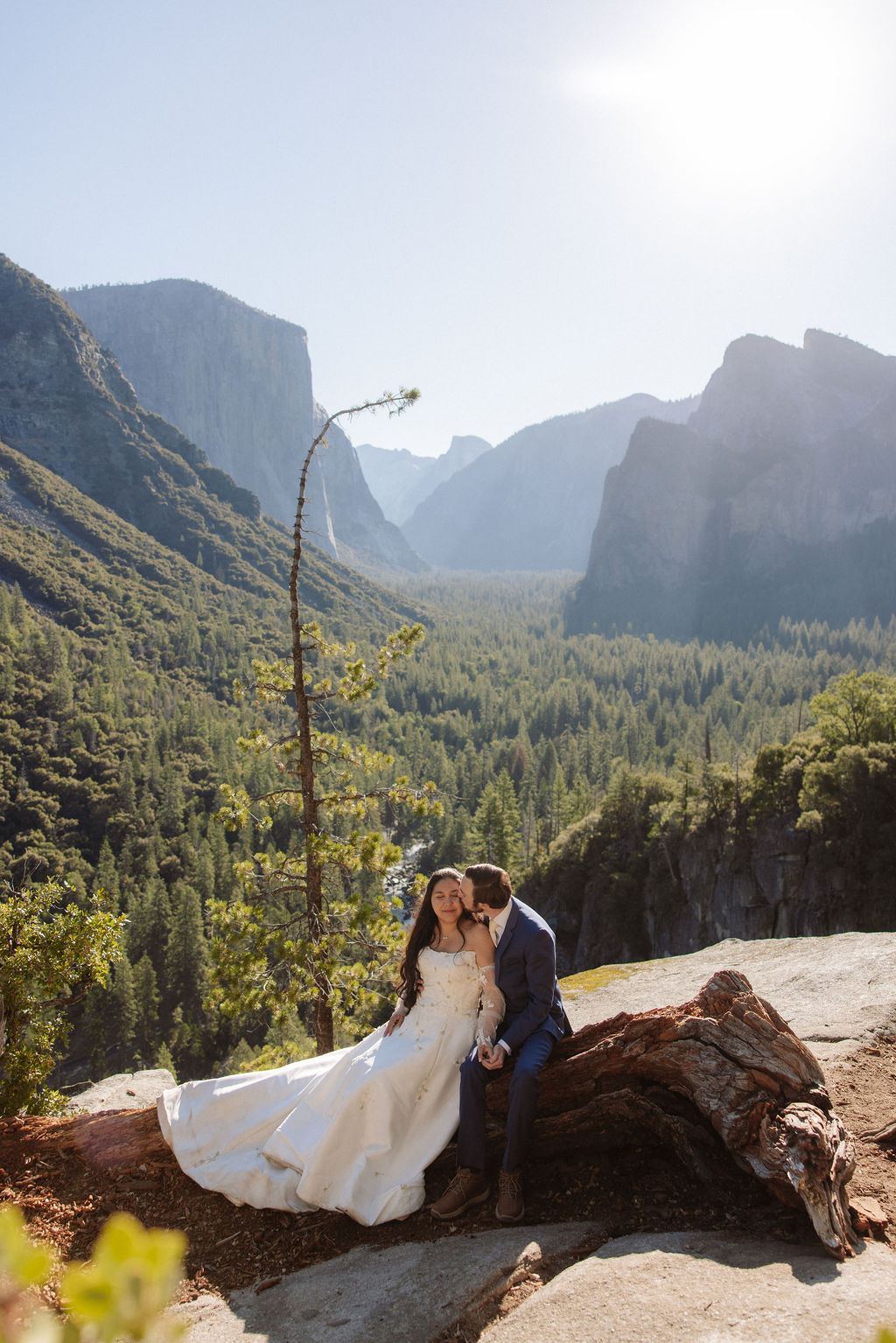 A bride and groom stand together on a rocky ledge overlooking a forested valley and mountains under a clear sky.