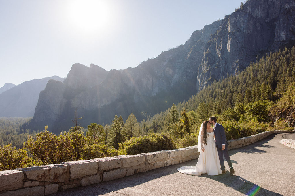 A bride and groom stand together on a rocky ledge overlooking a forested valley and mountains under a clear sky.