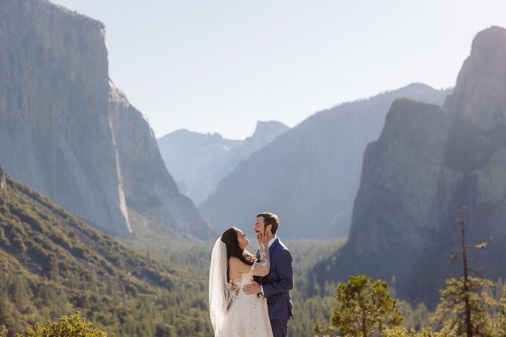 A bride and groom stand together on a rocky ledge overlooking a forested valley and mountains under a clear sky.