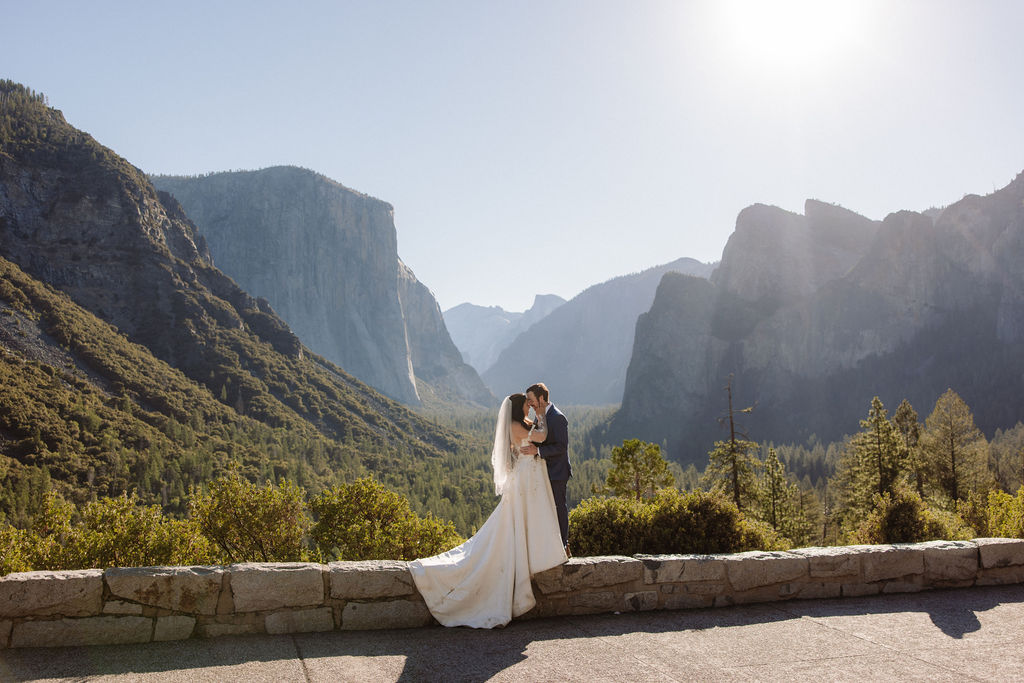 A bride and groom stand together on a rocky ledge overlooking a forested valley and mountains under a clear sky.