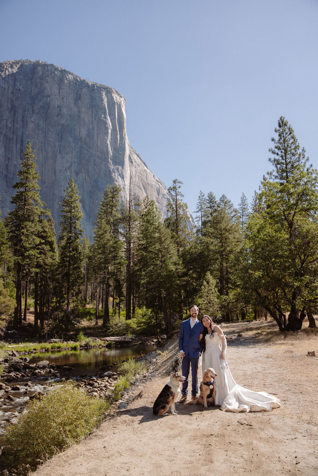 A bride and groom stand outdoors in front of a mountain and trees, accompanied by two dogs near a river on a sunny day.