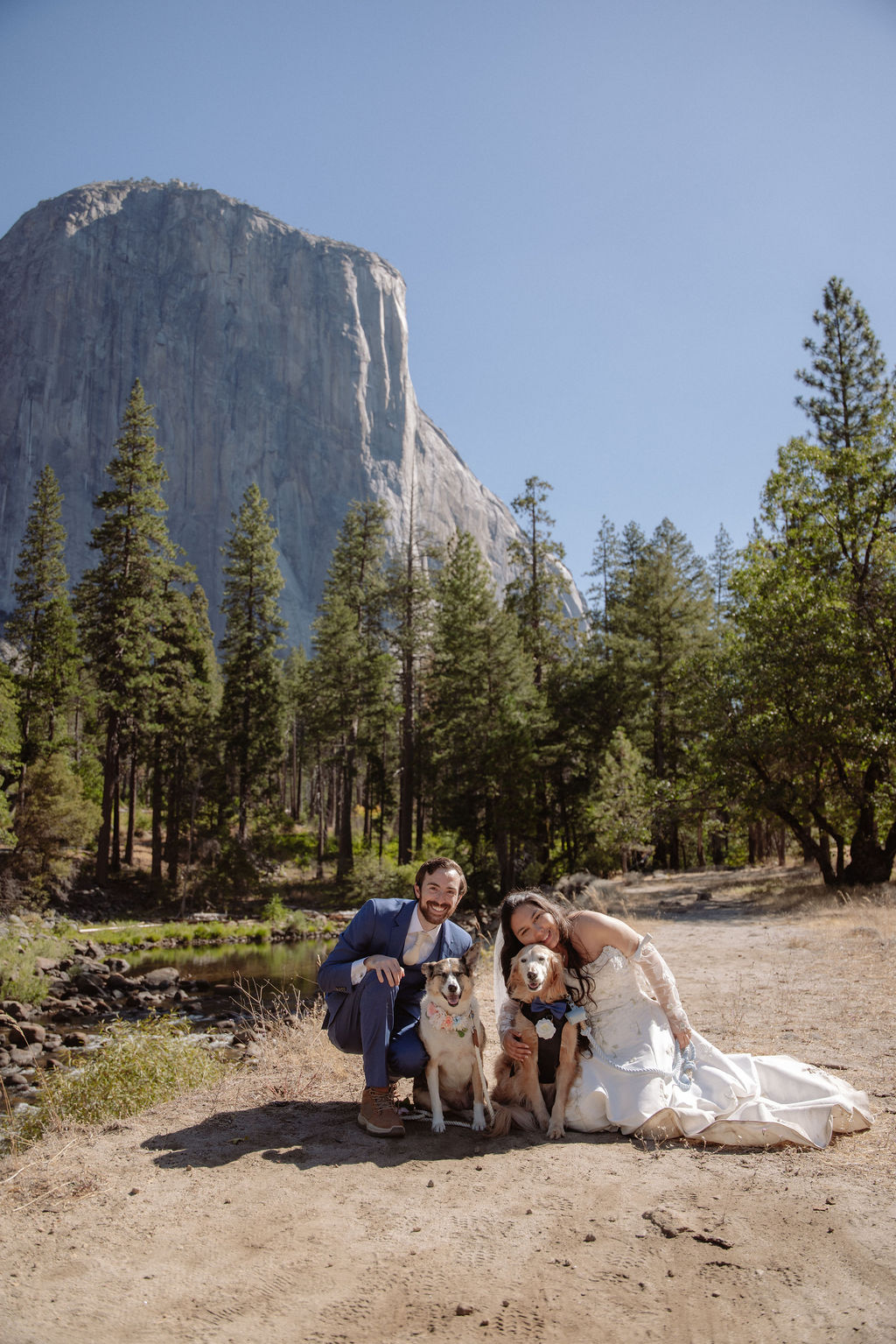 A bride and groom stand outdoors in front of a mountain and trees, accompanied by two dogs near a river on a sunny day.
