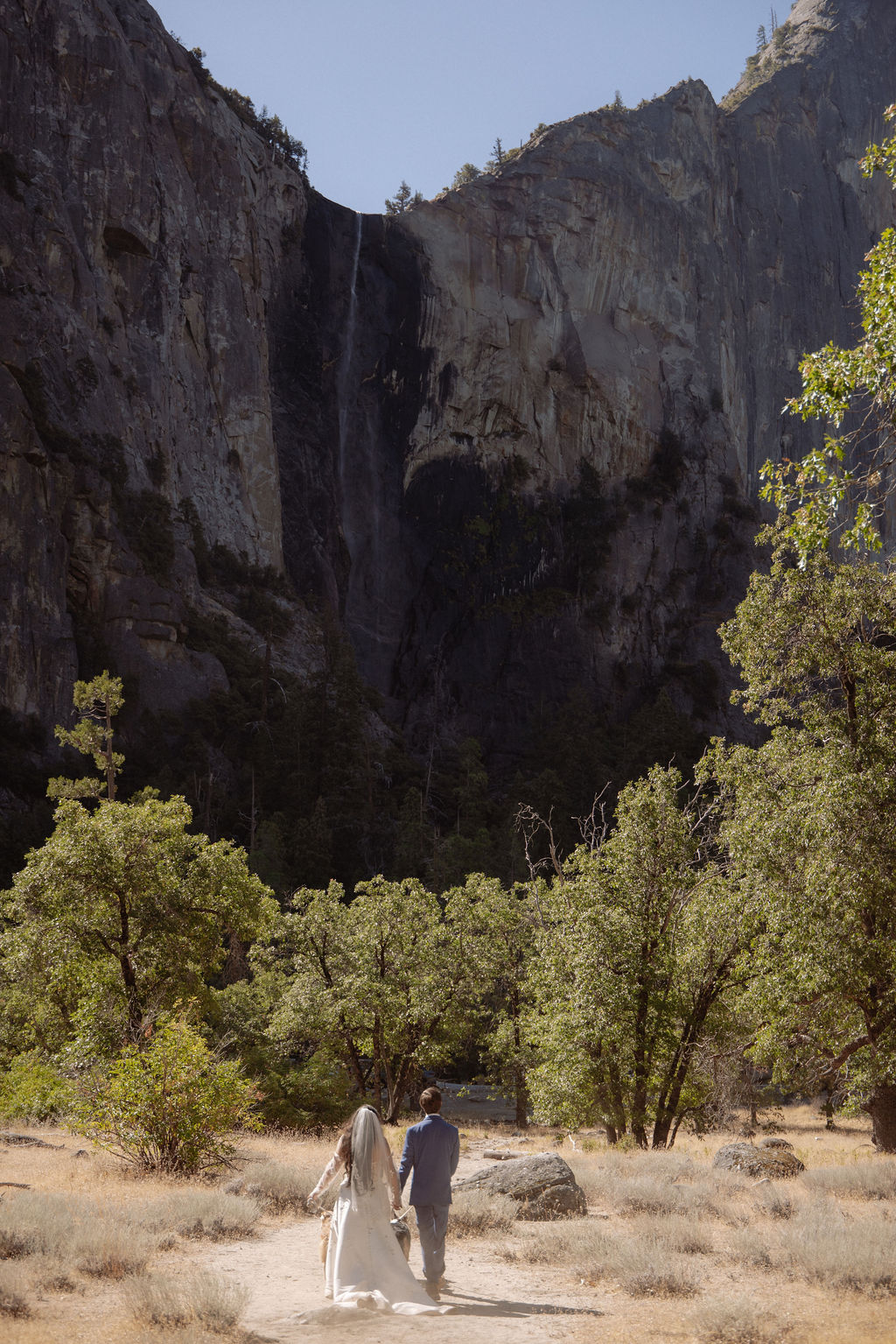 A couple holding hands walks along a dirt path through a grassy, tree-filled area with tall rocky cliffs and a waterfall in the background.