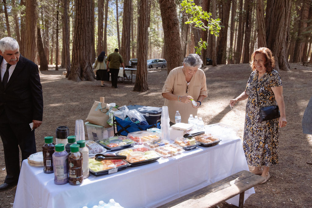 Three people stand by a picnic table covered with food and drinks in a wooded outdoor setting.