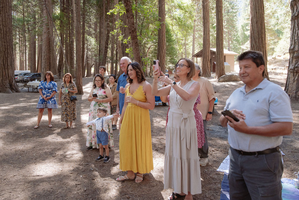 A group of people stand in a wooded outdoor area, some holding phones or cameras and watching something off-frame.