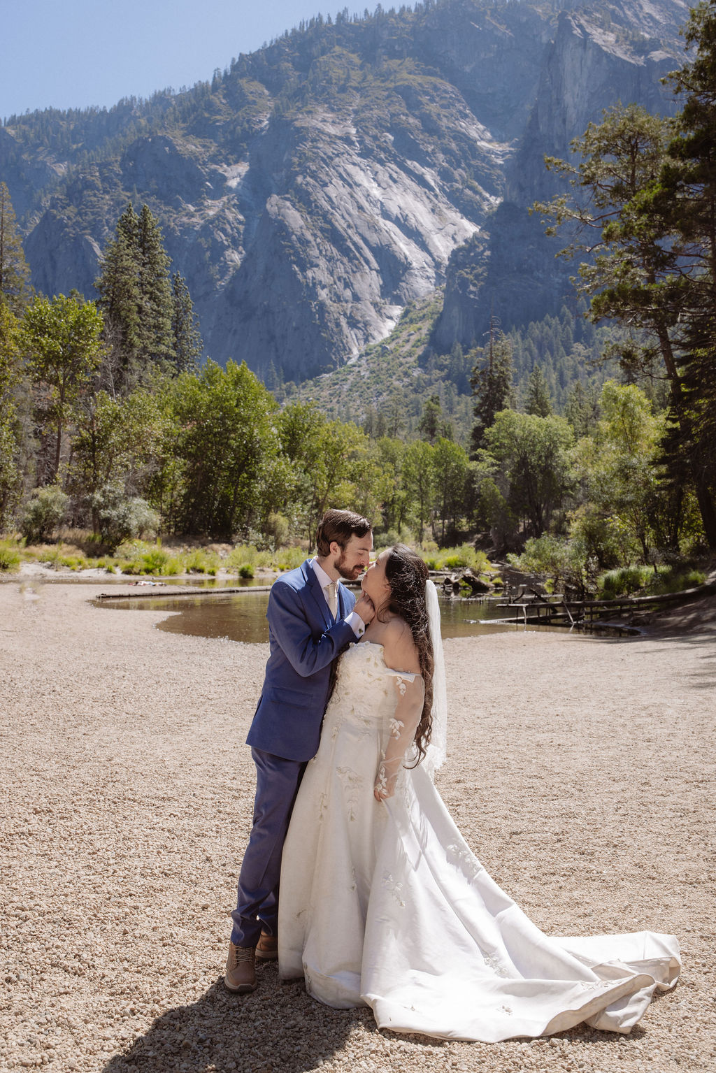A bride and groom stand together, holding hands and touching foreheads, on a sandy area with mountains and trees in the background for a wedding at Yosemite