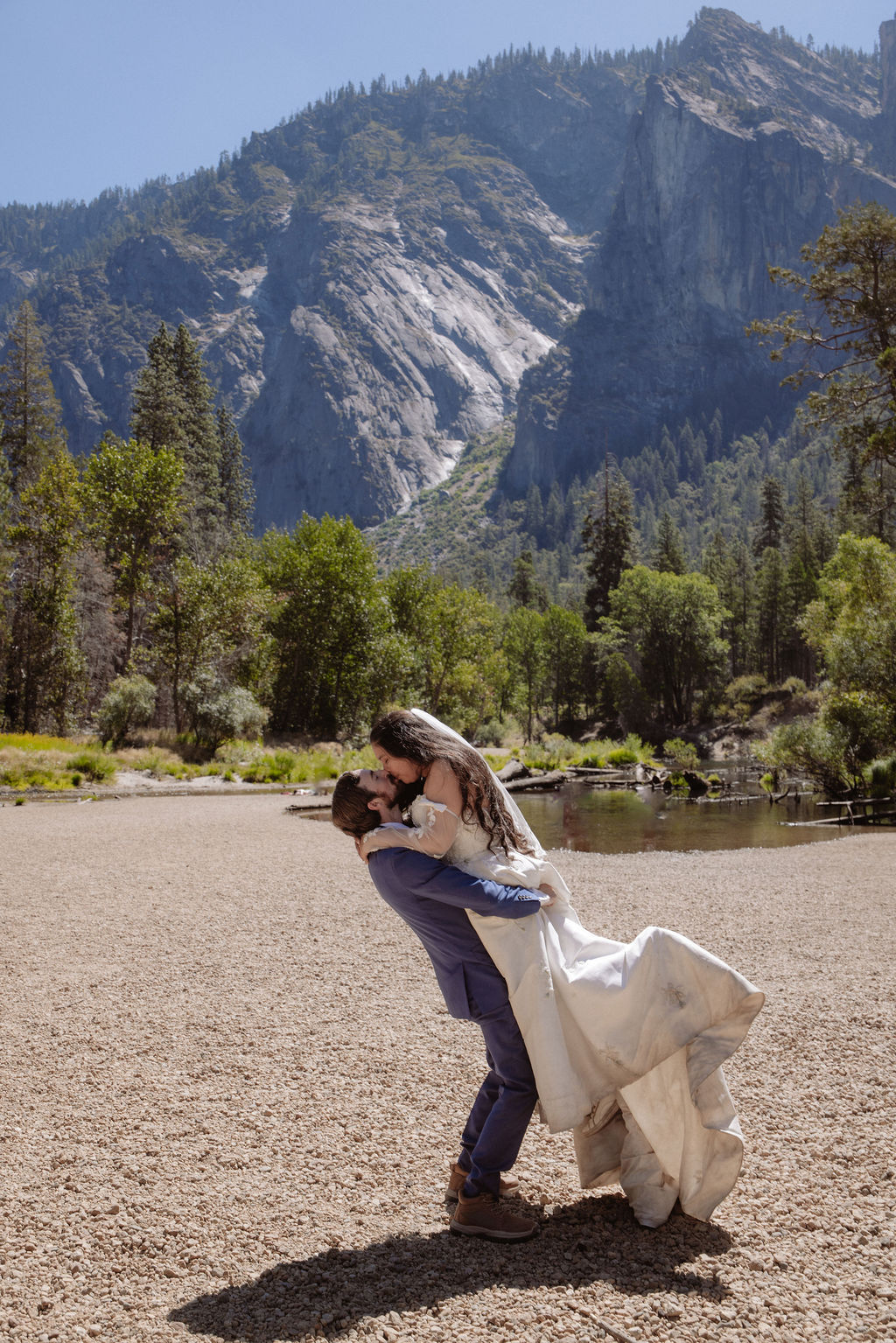 A bride and groom stand together, holding hands and touching foreheads, on a sandy area with mountains and trees in the background for a wedding at Yosemite