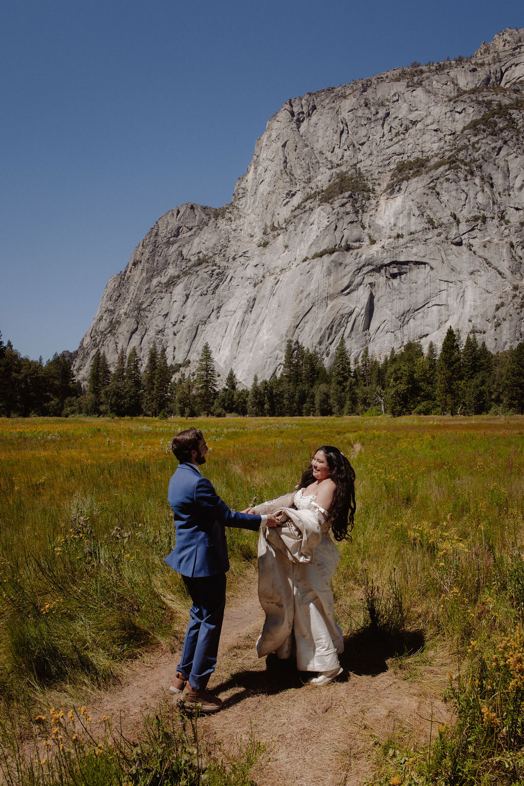 A bride and groom stand together, holding hands and touching foreheads, on a sandy area with mountains and trees in the background for a wedding at Yosemite
