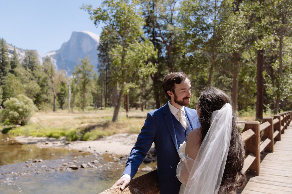 A bride and groom stand on a wooden bridge in a forested area, with mountains and trees in the background under a clear sky for a wedding at Yosemite