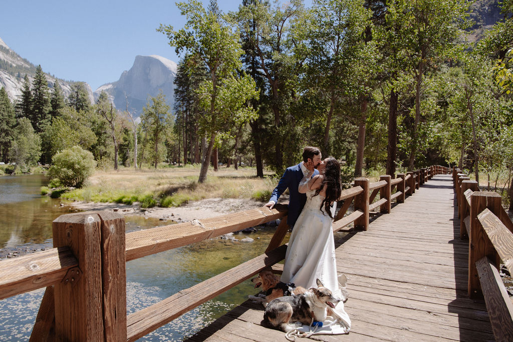 A bride and groom stand on a wooden bridge in a forested area, with mountains and trees in the background under a clear sky for a wedding at Yosemite