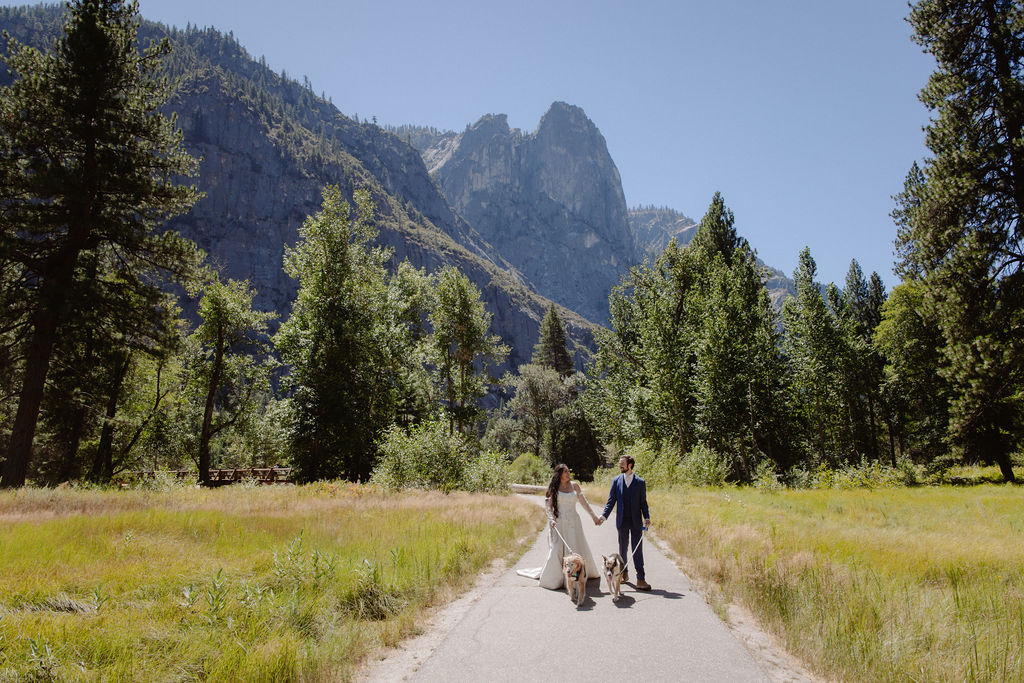 A bride and groom stand on a wooden bridge in a forested area, with mountains and trees in the background under a clear sky for a wedding at Yosemite