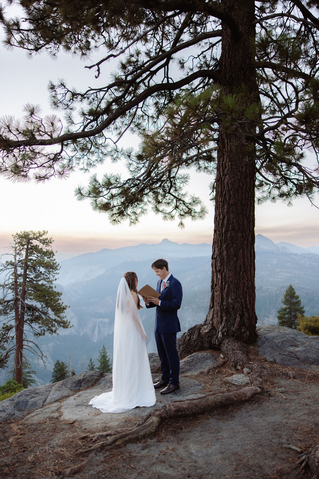 A bride in a white dress reads from a notebook to a groom in a navy suit during an outdoor wedding ceremony with mountains and trees in the background.