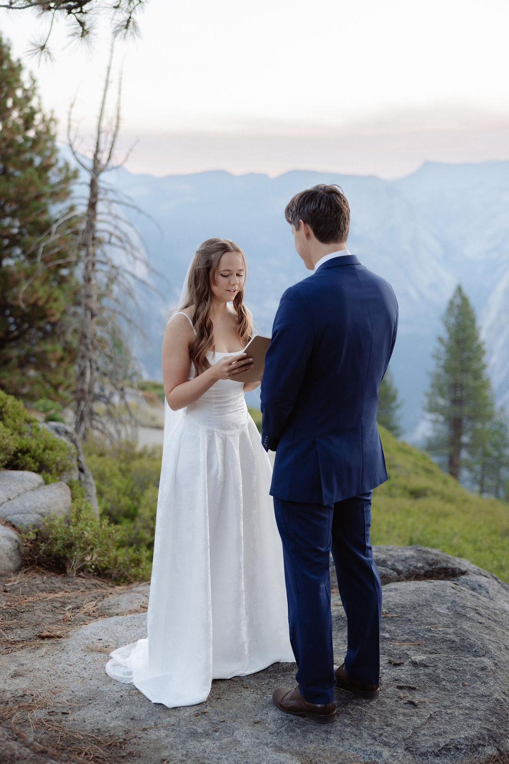 A bride in a white dress reads from a notebook to a groom in a navy suit during an outdoor wedding ceremony with mountains and trees in the background.