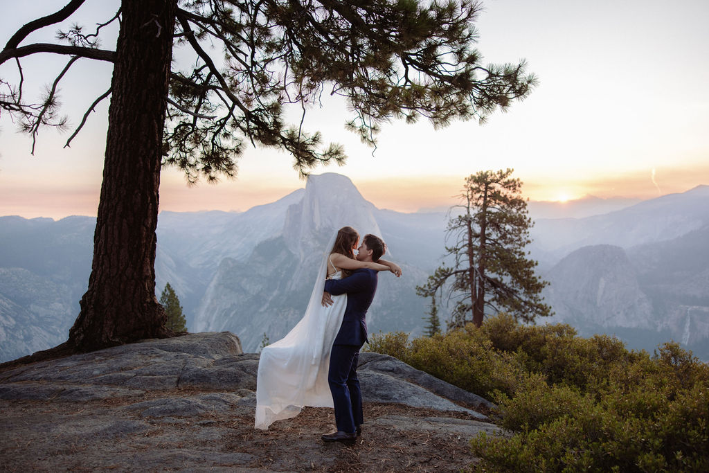A bride and groom embrace on a rocky ledge overlooking a mountain landscape at sunset, with trees and distant peaks visible in the background.