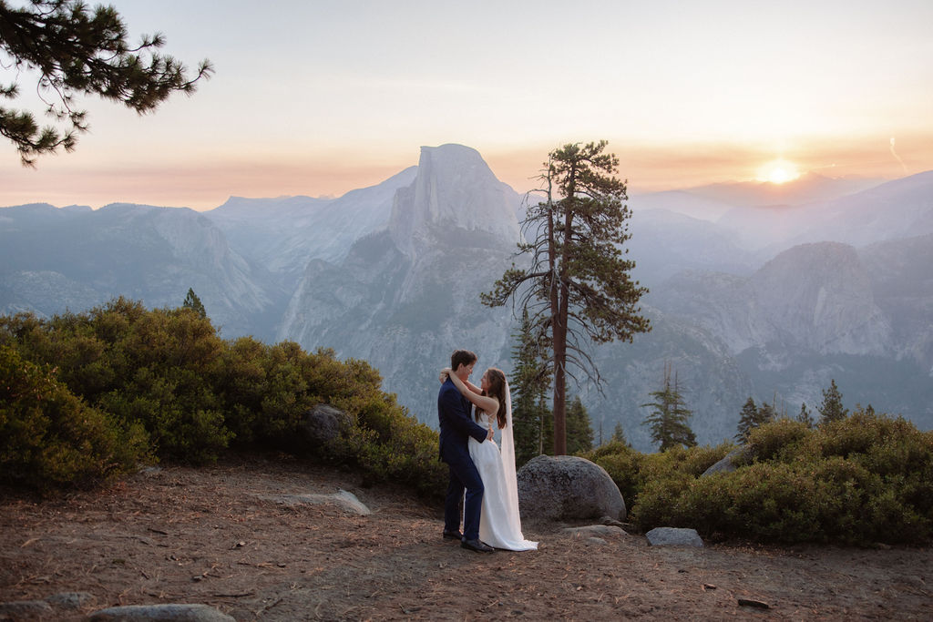 A bride and groom embrace on a rocky ledge overlooking a mountain landscape at sunset, with trees and distant peaks visible in the background.